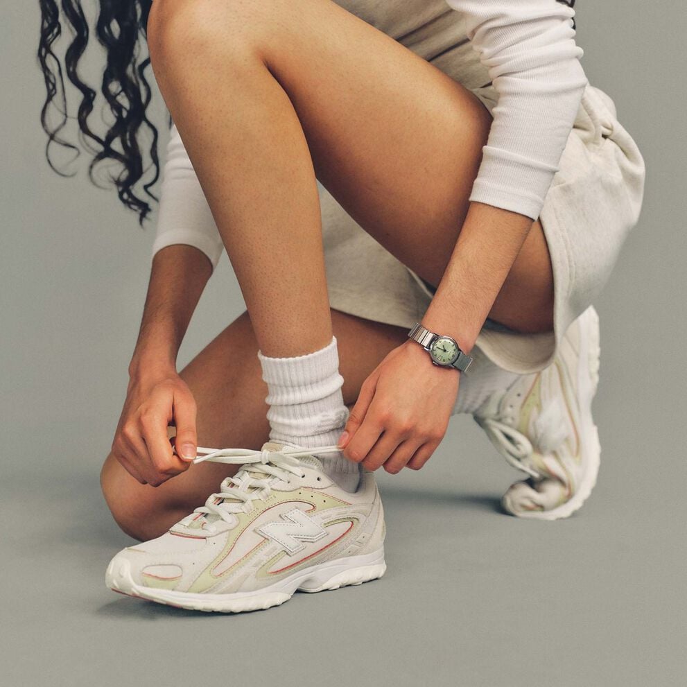 A person kneels on the ground while tying the laces of a white New Balance sneaker with pastel accents.